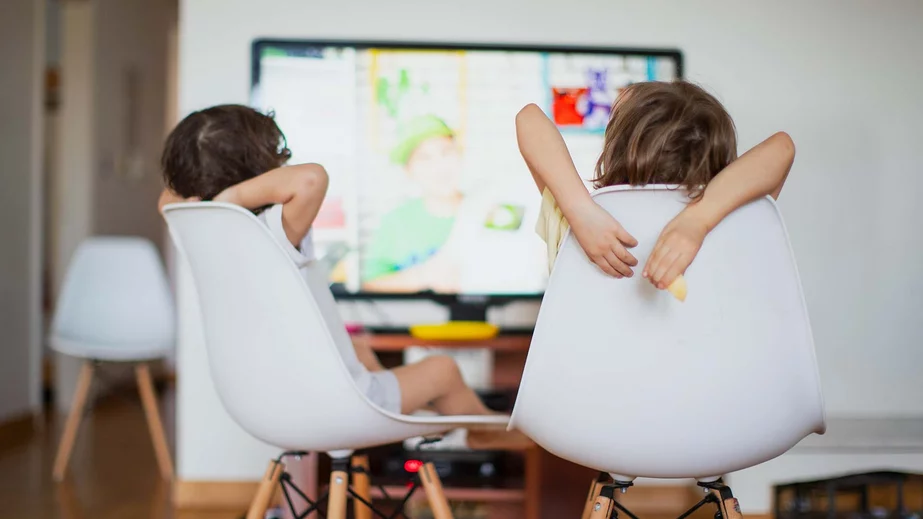 Zwei kleinere Kinder sitzen in gemütlicher Pose mit nach hinten verschränkten Armen auf weißen Stühlen mit vollflächiger und geschwungener Rückenlehne und schauen auf den Bildschirm eines Fernseher an einer Wand im Hintergrund, der den Verbrauch von Kilowattsunden symbolisiert.