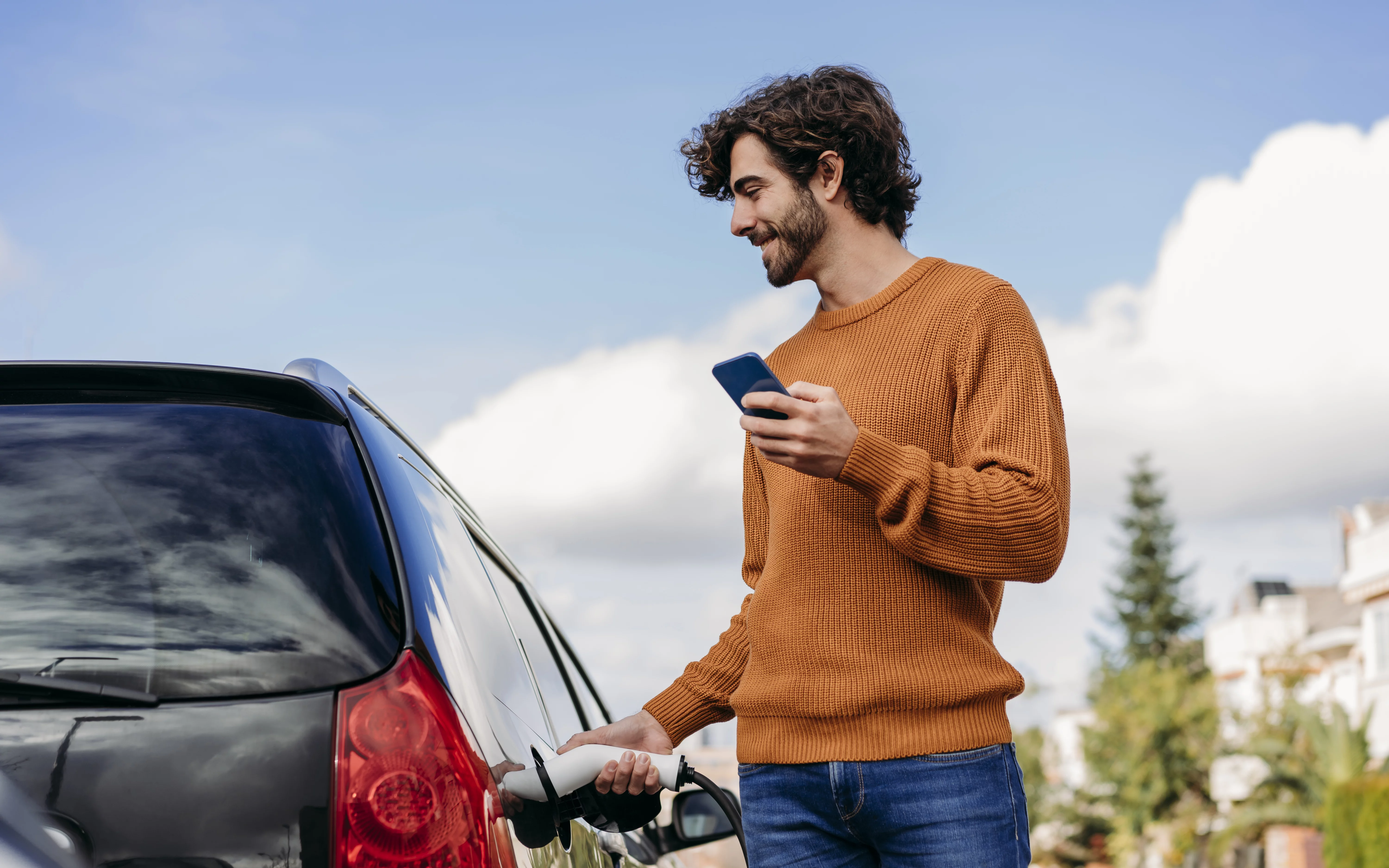 Mann steht mit Smartphone an seinem Auto und hat den Ladestecker in der Hand