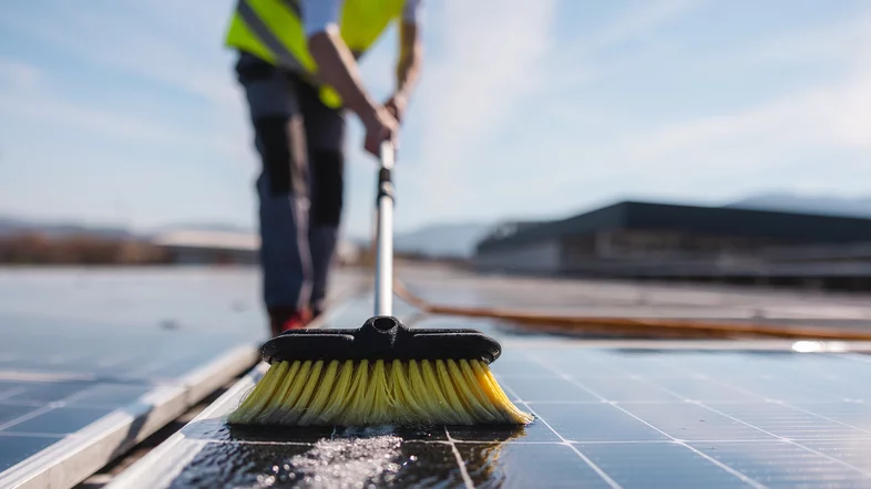 Eine Person, die zwischen zwei Solar-Paneelen einer Photovoltaikanlage steht, sorgt mit einem Besen für die Reinigung der Solaranlage und entfernt Verschmutzungen.